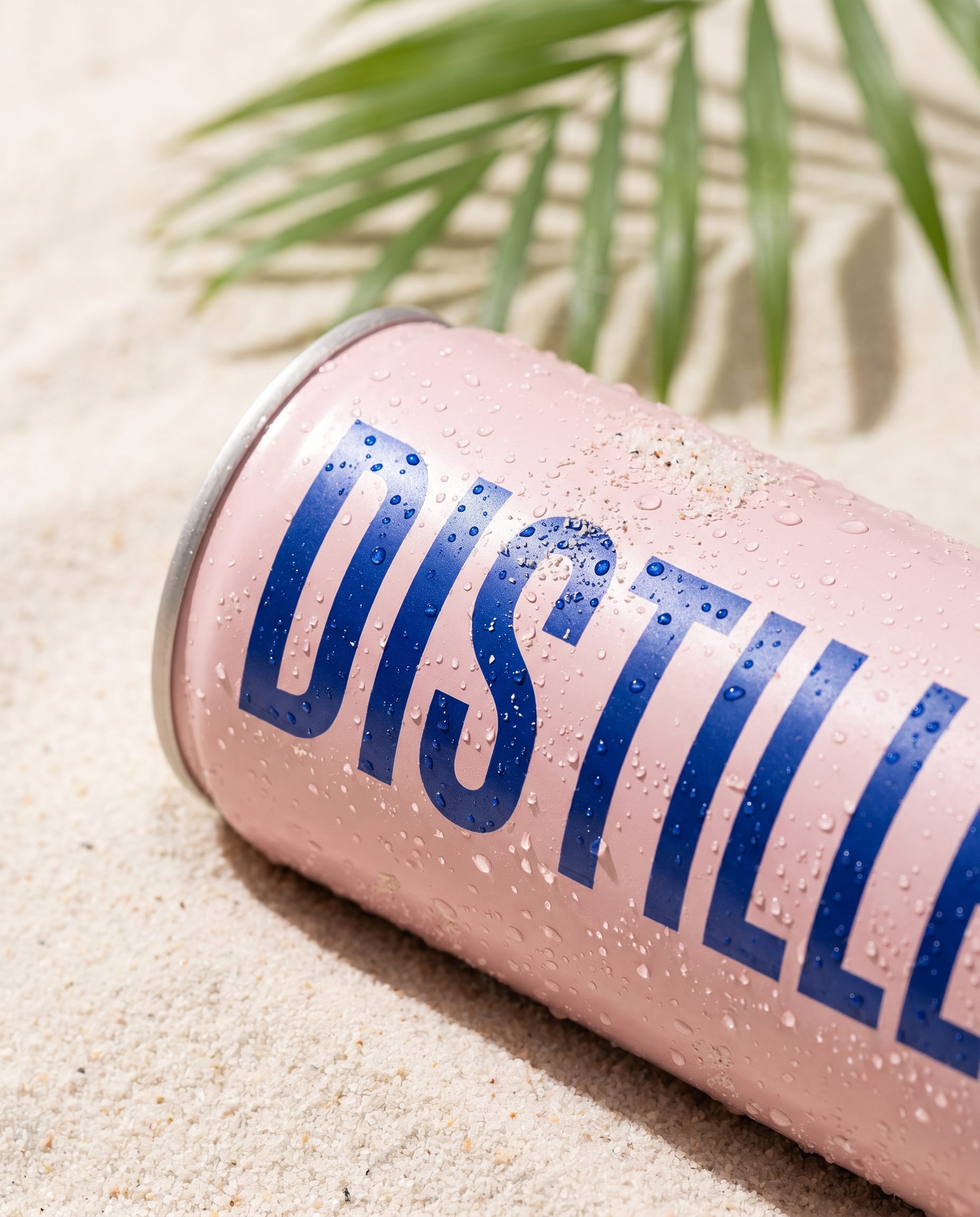 A macro of the chilled can resting on pale sand with a crisp palm-leaf shadow