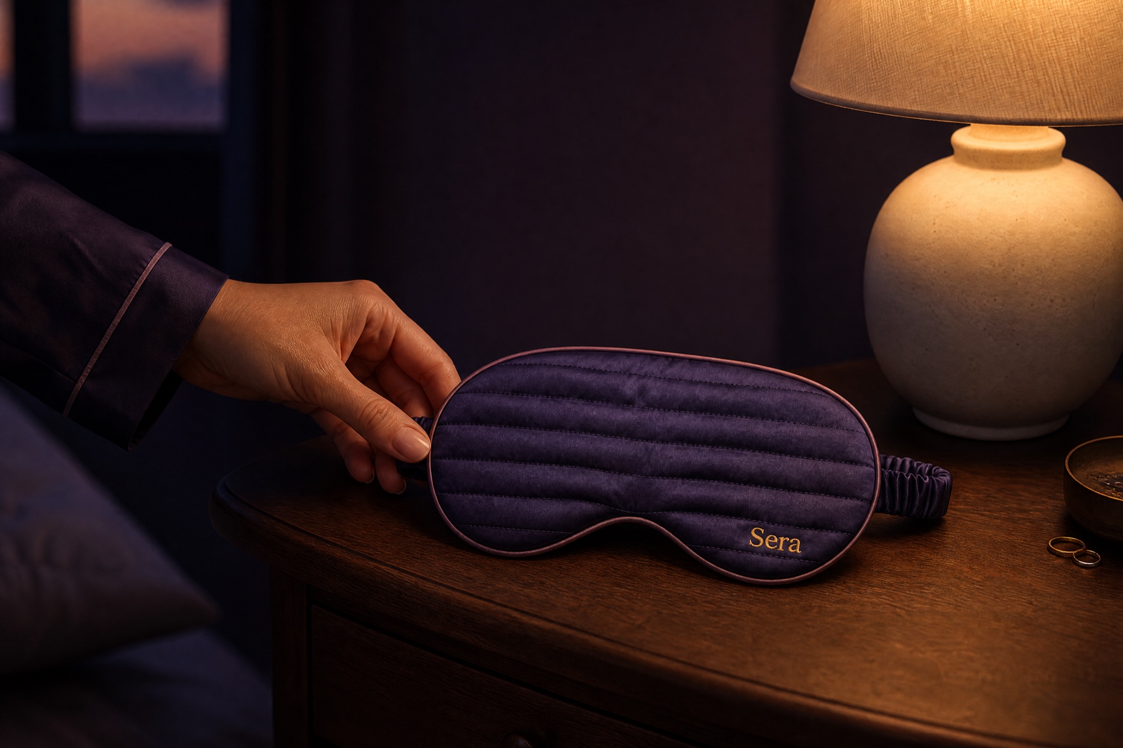 A hand setting the mask on a wooden nightstand beside a glowing lamp at dusk