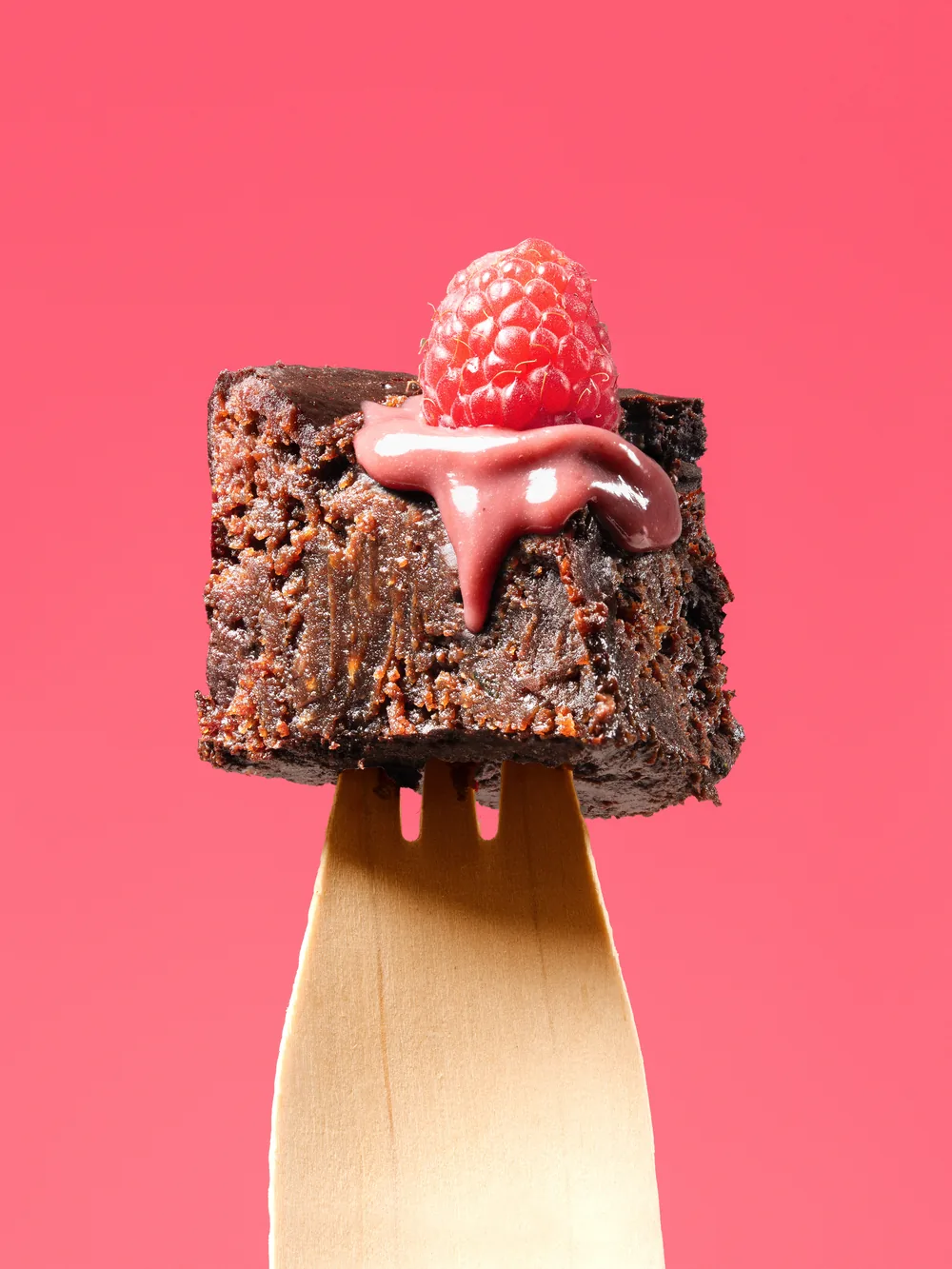 Square of chocolate brownie on a wooden fork, topped with a raspberry and pink glaze.