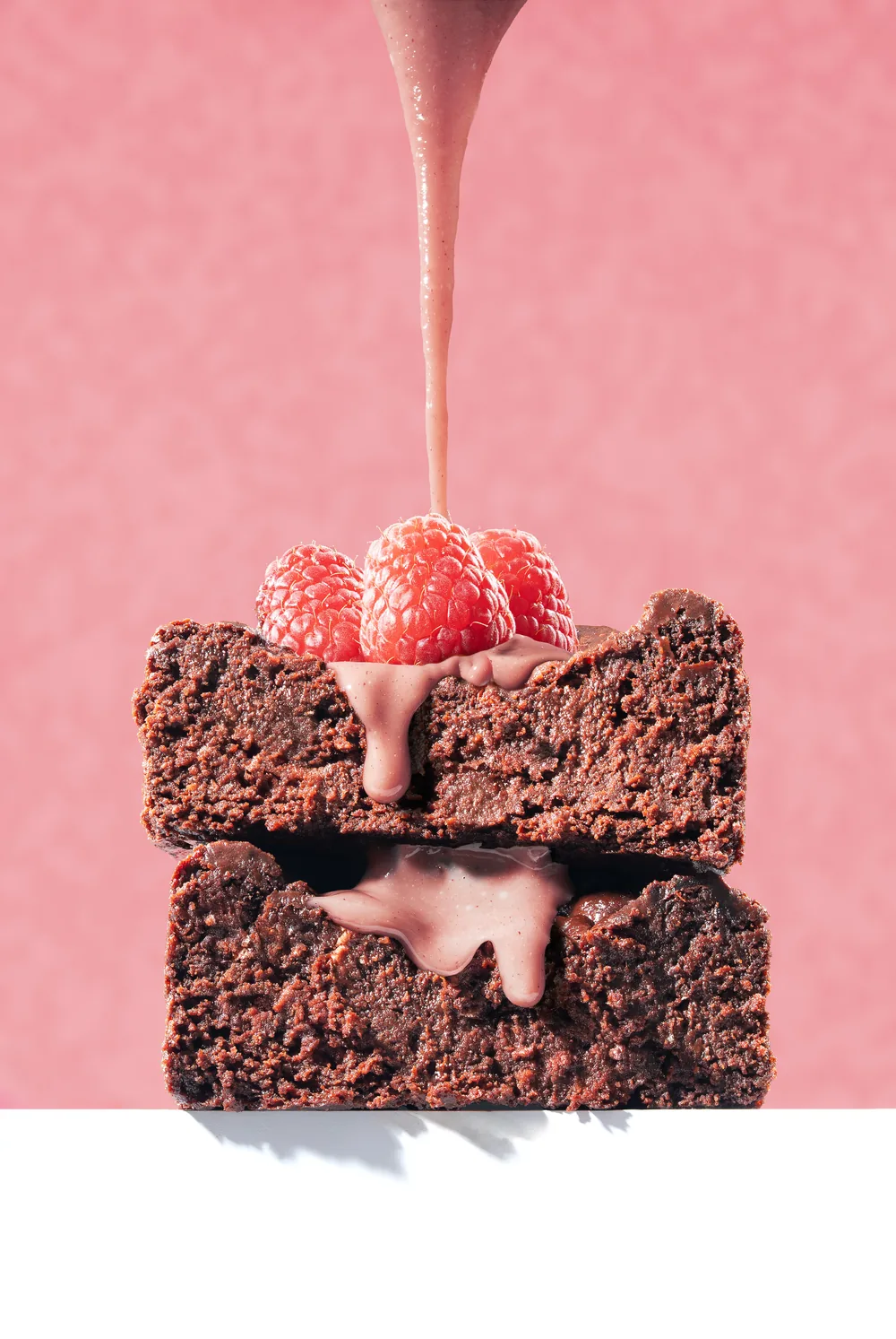 Stack of chocolate brownies topped with raspberries as pink syrup is poured over them.