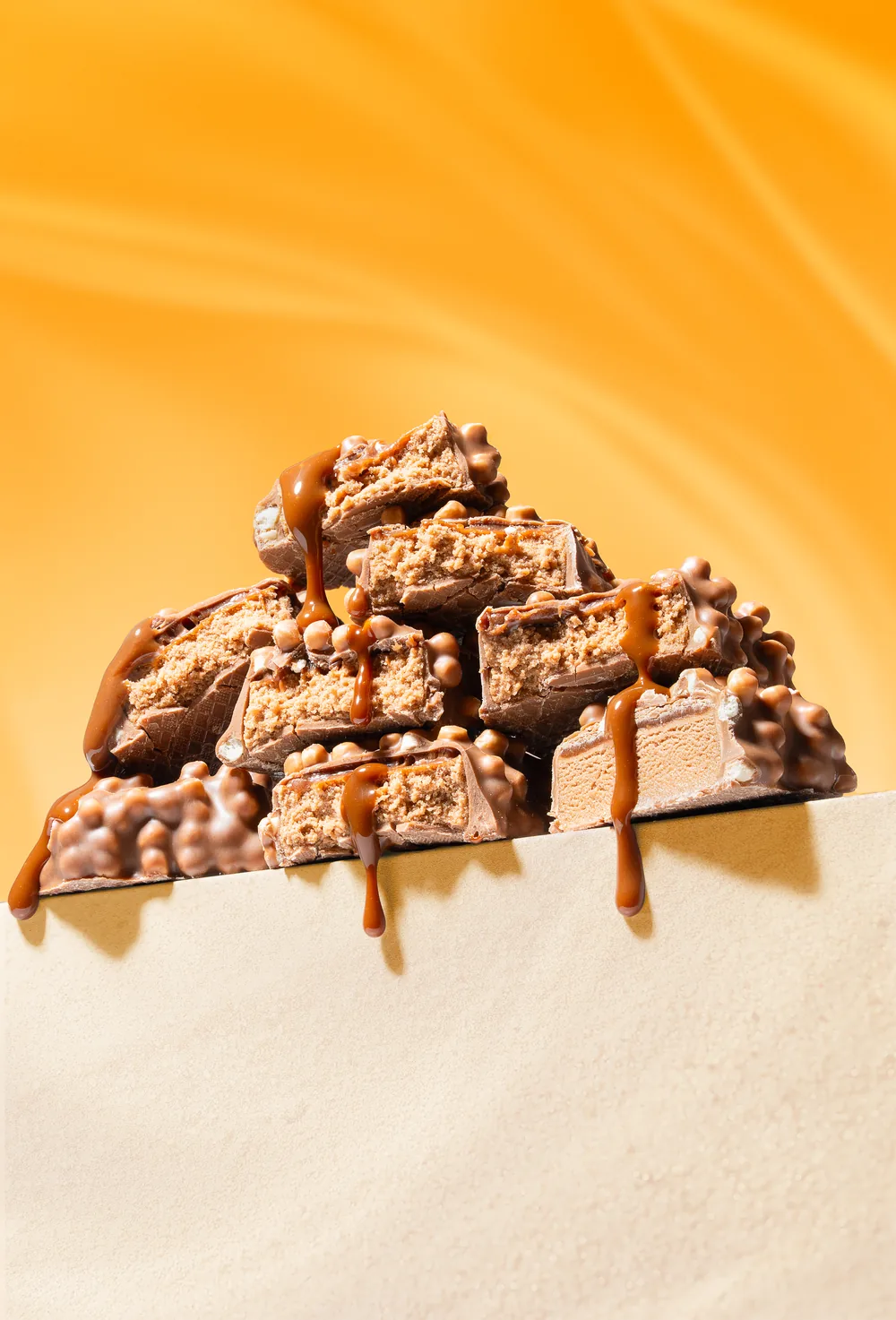 Stack of chocolate and peanut candy bars with caramel drizzle on a bright orange background.