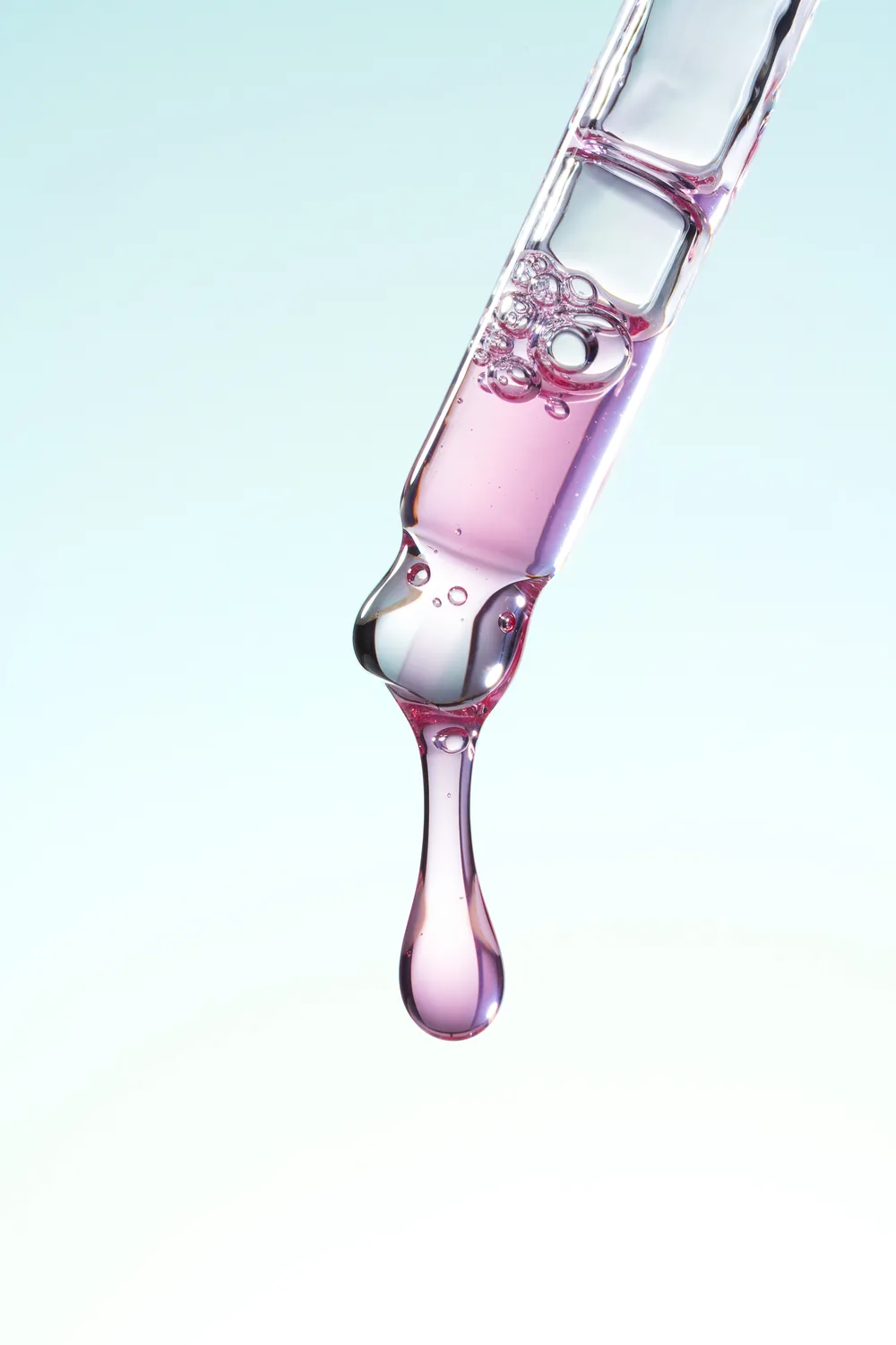 Glass pipette releasing a drop of pink serum against a pale blue background.