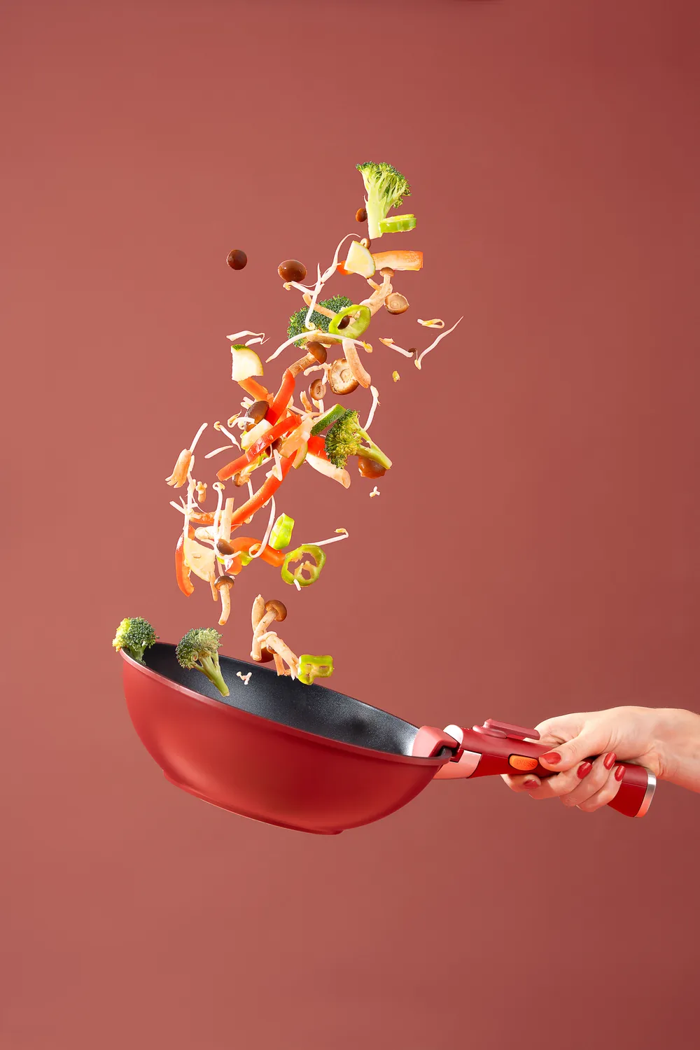 Chopped vegetables flying out of a red frying pan against a red background.