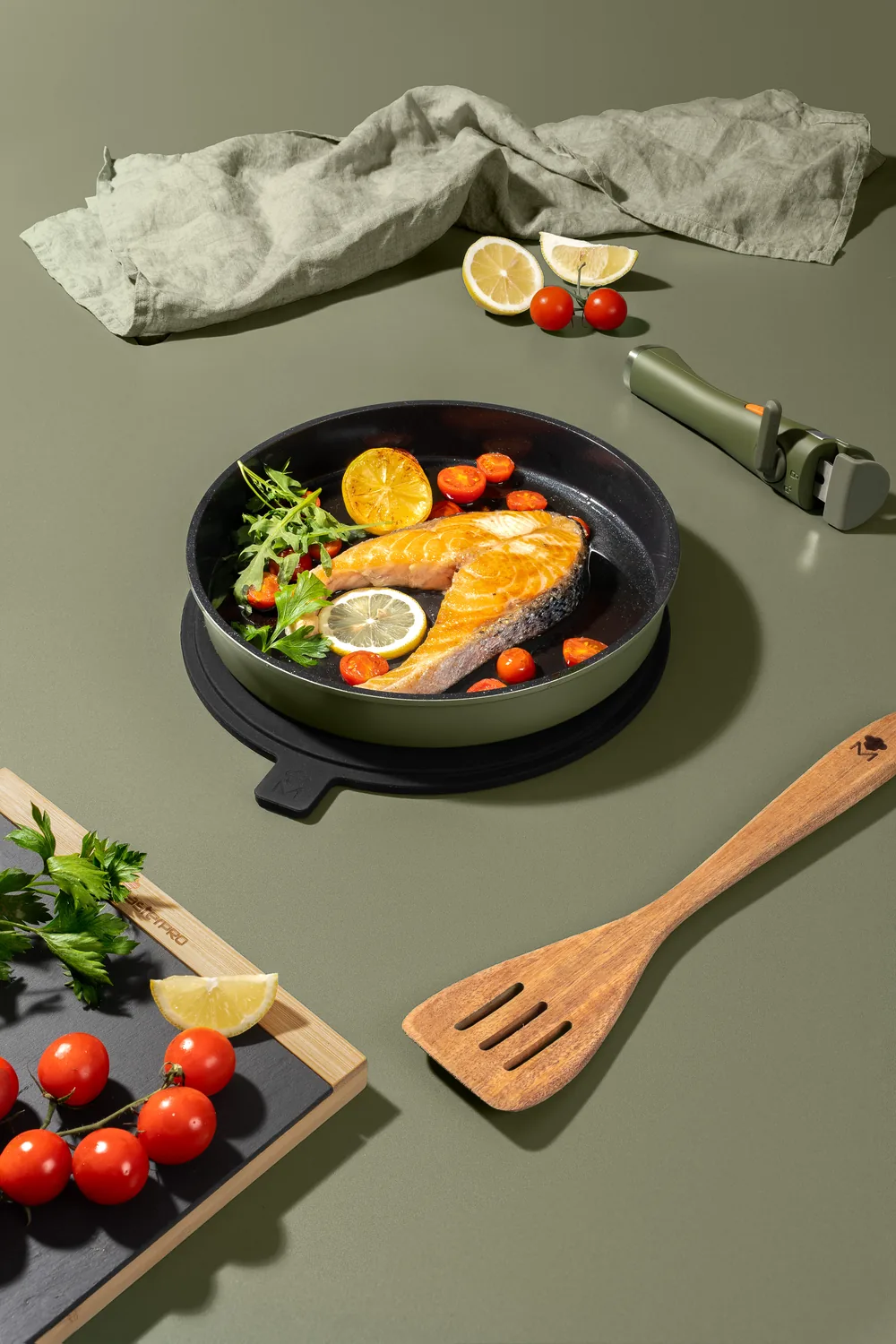 Salmon fillets in a MasterPro skillet with lemon, tomatoes and herbs on a sage green table.