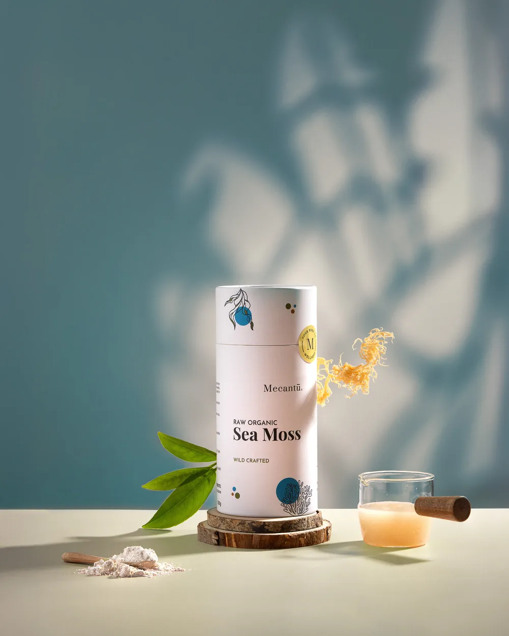 Mecantu Sea Moss jar on a pale surface with leaves, a wooden scoop and a glass of tonic.