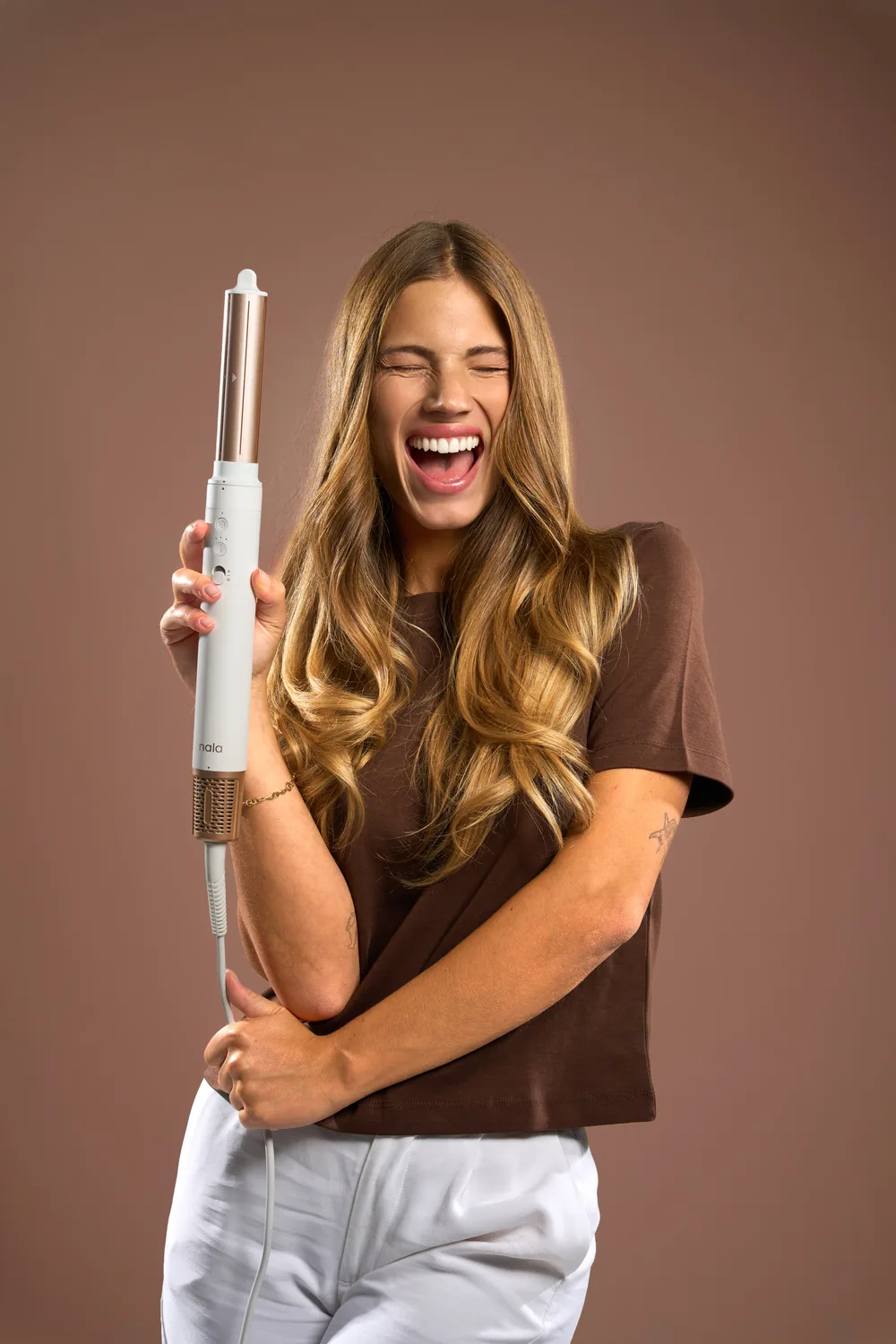 Woman laughing while holding a cordless Nala hair styler against a warm brown background.