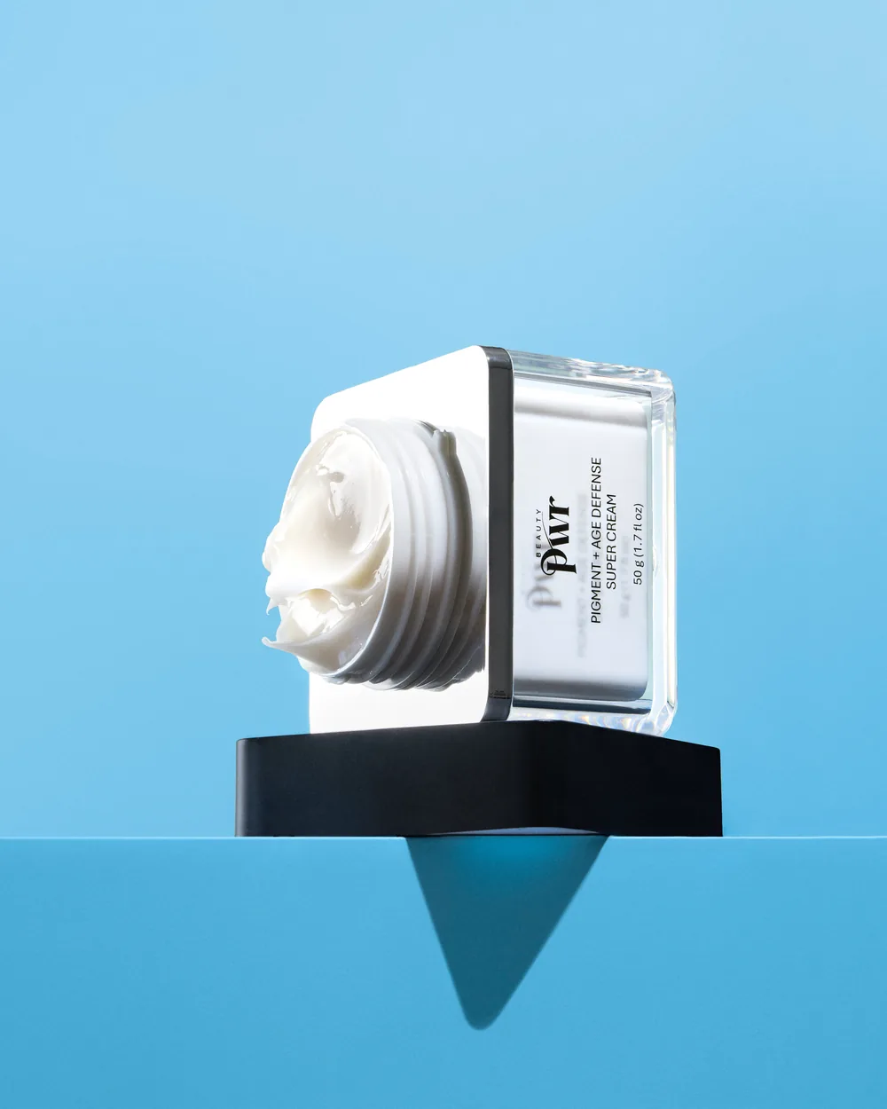 Open PWR Beauty Pigment + Age Defense Super Cream jar tilted on a black stand over a sky-blue ledge.