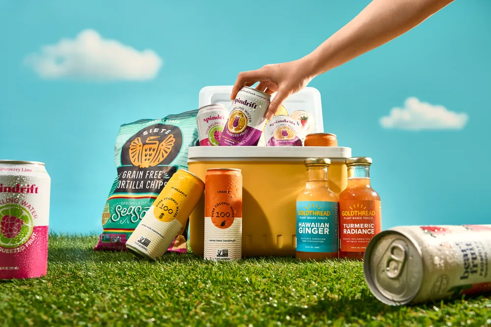 Hand lifting a drink from a cooler on grass, surrounded by Simply Real Market cans and snacks.