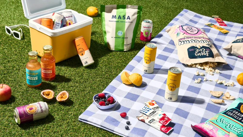 Picnic blanket on grass laid with Simply Real Market snacks, drinks and fresh fruit.