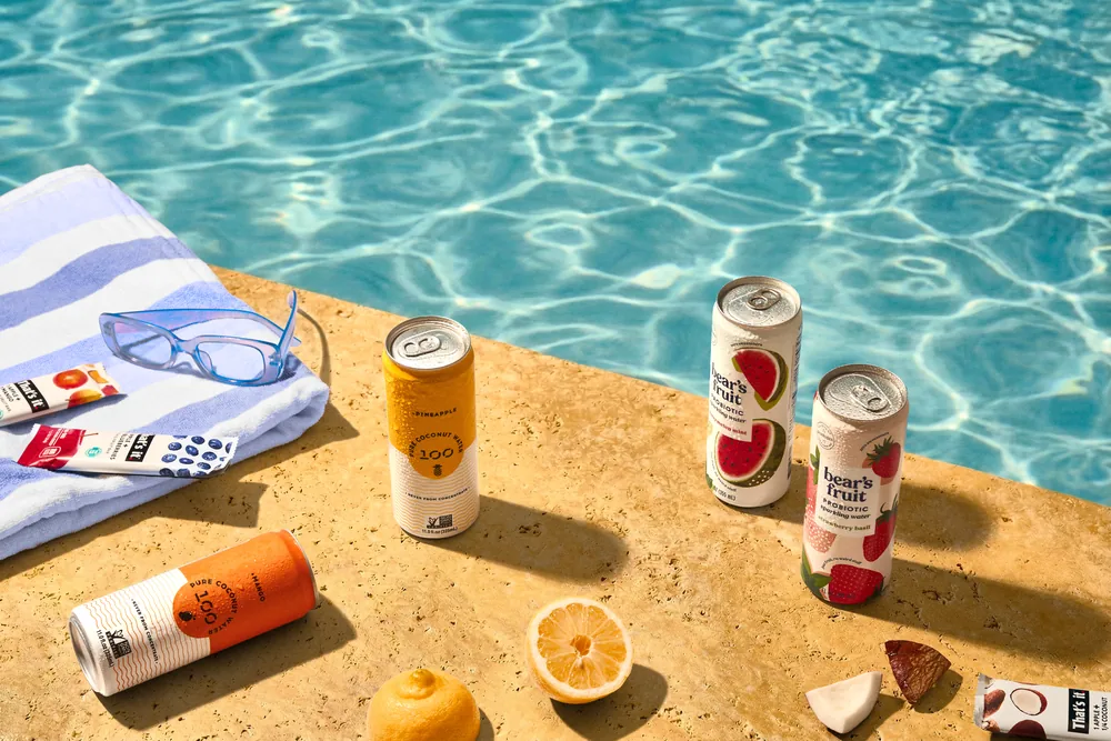 Chilled cans and snacks arranged on a sunlit pool ledge beside a folded towel.