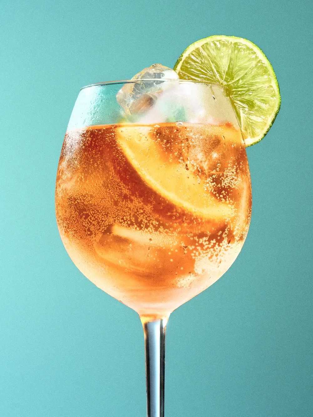 Spritz cocktail in a stemmed glass with an orange slice and lime wheel against a teal background.