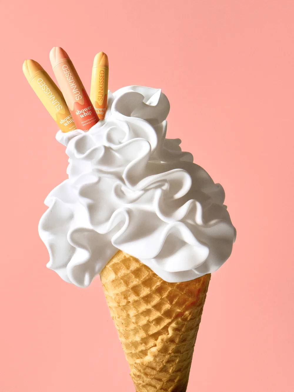 Sunkissed Shower Whip tubes topping a swirl of white whipped foam in a waffle cone against a peach background.