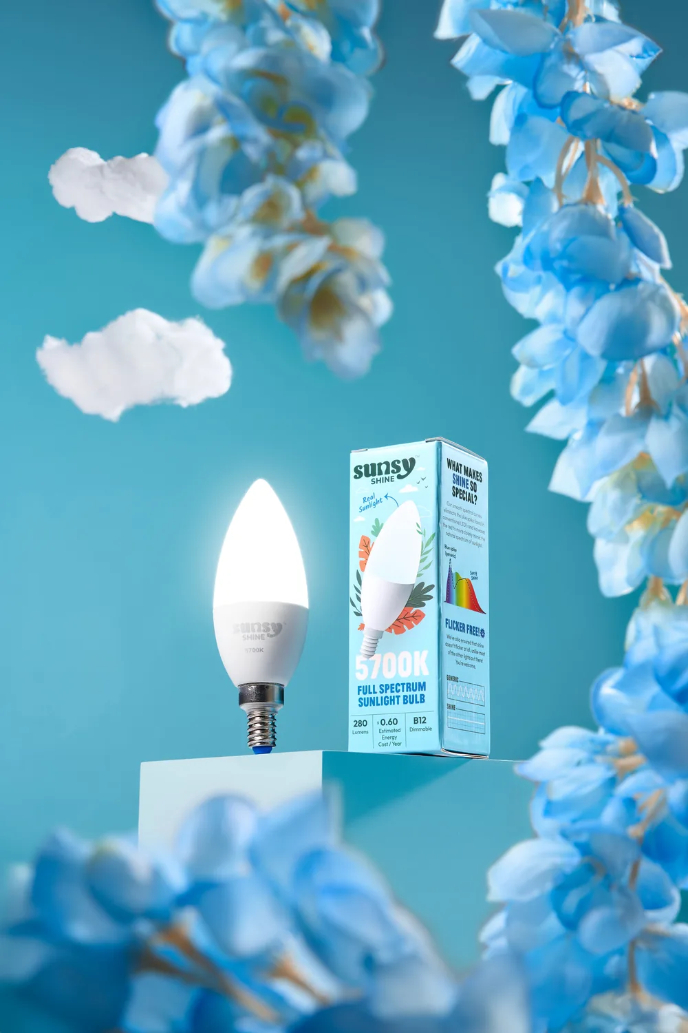 Sunsy full spectrum daylight bulb lit on a pedestal, framed by blue delphinium flowers.